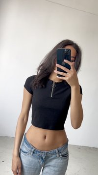 Černý žebrovaný crop top s předním zipem a kulatým táhlem, v kombinaci se světle modrými džíny. Minimalistický design, jednobarevné pozadí.