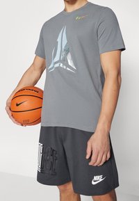 Hombre vistiendo una camiseta Nike gris y pantalones cortos Nike negros sosteniendo un balón de baloncesto naranja frente a un fondo liso.
