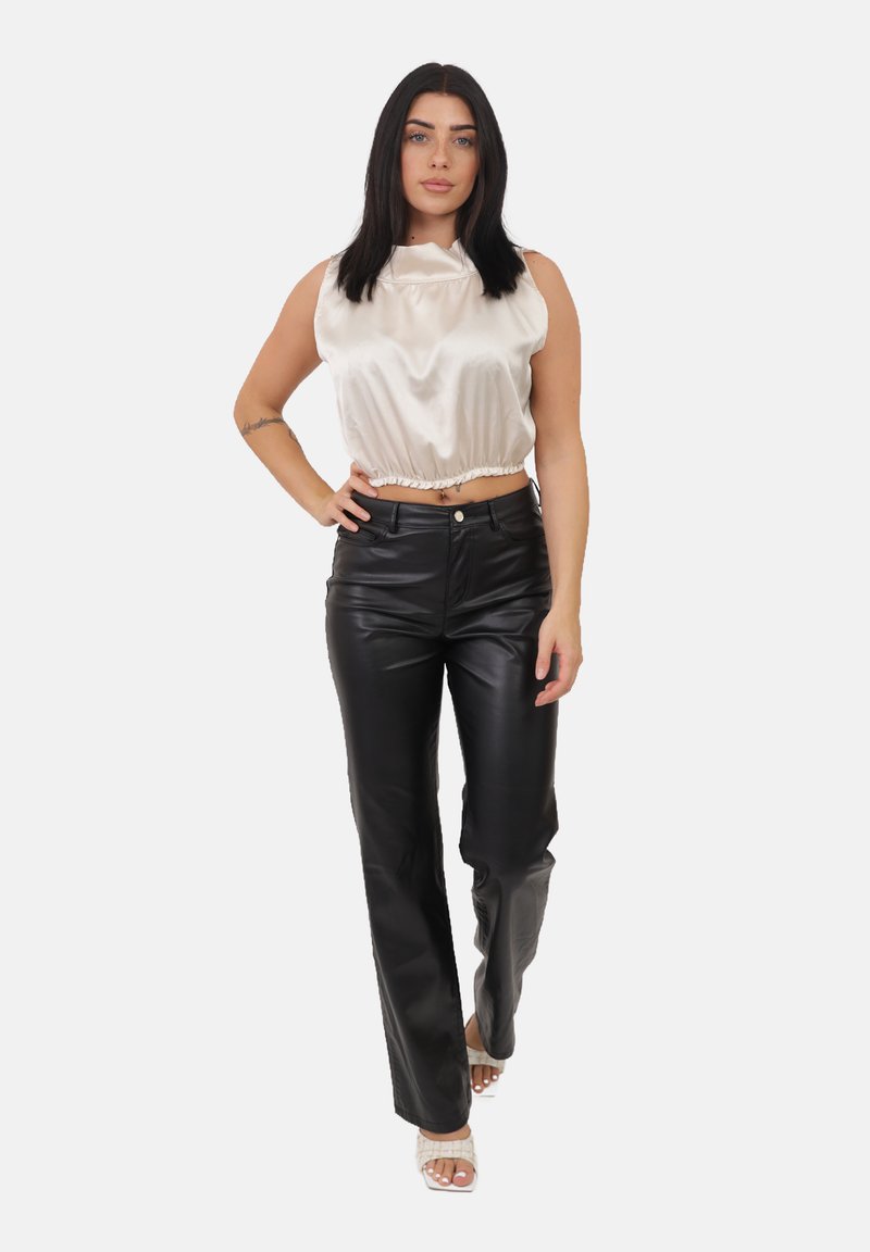 Donna con capelli scuri che indossa un top corto in satin avorio senza maniche, pantaloni di pelle nera e sandali col tacco bianchi, in piedi su uno sfondo bianco.