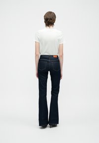 Persoon met kort haar in een wit T-shirt, donkere altaar-high-waisted flared jeans en zwarte schoenen, die staat met de rug naar een effen witte achtergrond.