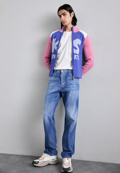 Farbenfrohe Jacke mit blauem Körper und pinken Ärmeln, weißen Akzenten und "SPORT"-Schriftzug. Hellblaue Jeans und mehrfarbige sportliche Sneaker.