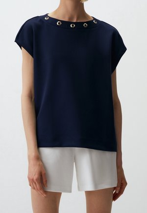 Woman wearing a navy blue short-sleeve top with metal grommets at the neckline and white knee-length shorts against a plain background.