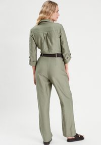 Khaki-Overall mit Kragen, Knopfmanschetten und gerafftem Rücken. Er besteht aus einem strukturierten Stoff und hat eine weite Beinform, kombiniert mit schwarzen Sandalen.