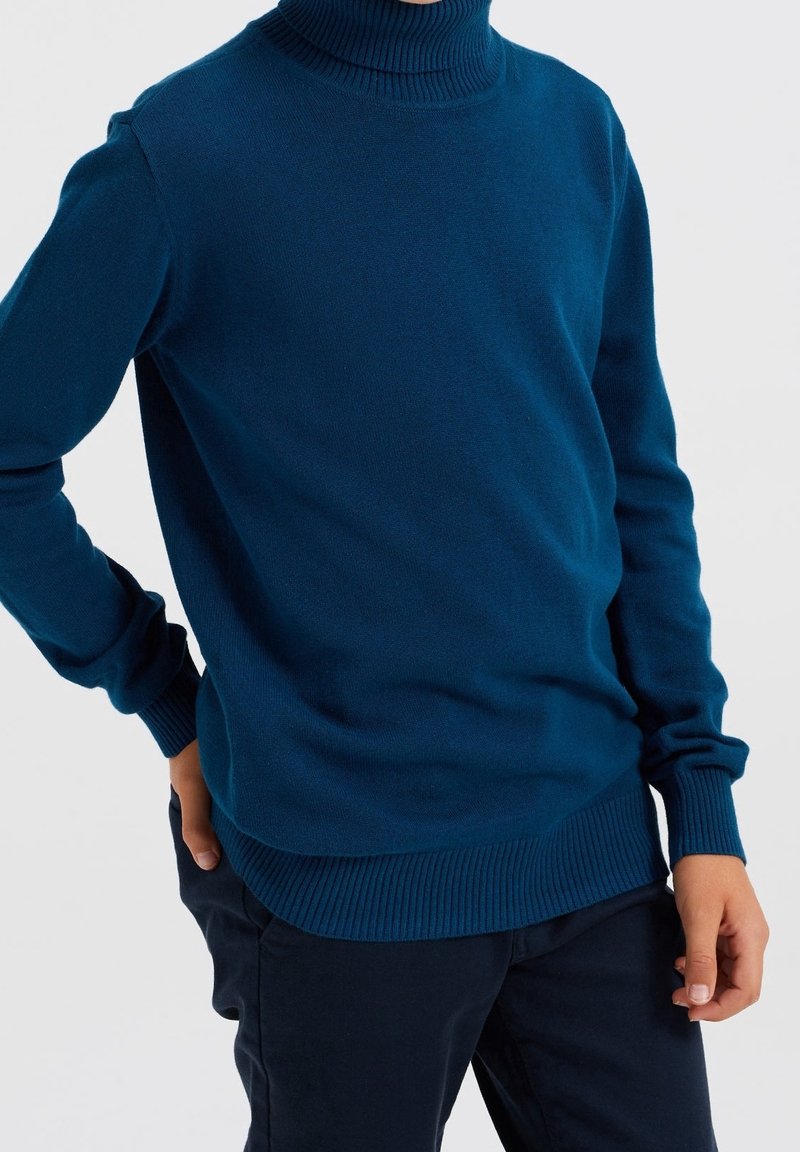 Personne portant un pull col roulé ajusté, manches longues, bleu foncé, avec des poignets et un ourlet côtelés, associé à un pantalon foncé.