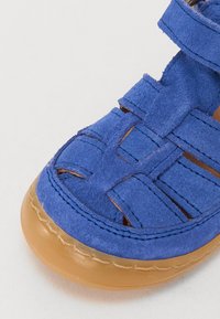 Sandal in suede bluè con design intrecciato, dotato di sezioni a taglio, punta rotonda e suola in gomma beige. I dettagli di cucitura sono visibili.