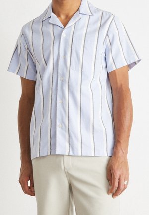Homme portant une chemise bleu clair à manches courtes boutonnée avec des rayures verticales blanches et grises et un pantalon clair.
