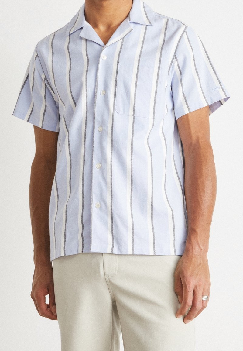 Homme portant une chemise bleu clair à manches courtes boutonnée avec des rayures verticales blanches et grises et un pantalon clair.
