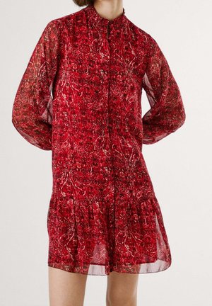 Robe rouge translucide à manches longues avec des motifs cachemire complexes et un volants froncés près de l'ourlet, portée par une personne debout avec les mains derrière le dos.