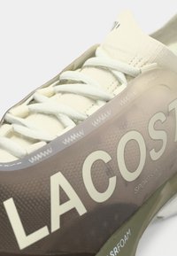 Sneaker Lacoste με σχέδιο σε καφέ και κρέμα, υφή στο επάνω μέρος, επίπεδα κορδόνια και λεπτομέρειες λογότυπου. Κατασκευασμένο από ελαφριά υλικά και με ορατό πάτο από αφρώδες υλικό.