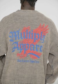 Pletený sveter s rebrovanou textúrou, so širokým grafickým motívom v modrej a červenej farbe, na ktorom je napísané "Multiply Apparel" s dizajnom krídel.