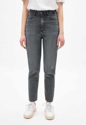 Osoba nosící vybledlé černé džíny s vysokým pasem a bílé tenisky, stojící s uvolněnýma rukama podél těla na jednobarevném bílém pozadí.