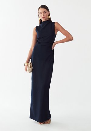 Donna che indossa un abito lungo blu navy senza maniche, che tiene una pochette intrecciata, con orecchini, bracciale e sandali dorati su sfondo bianco.