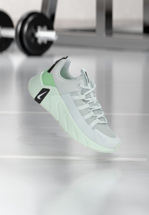 Hellgrauer und mintgrüner Nike-Sneaker mit klobiger Sohle, Mesh-Obermaterial und sichtbaren Schnürsenkeln, gezeigt auf einem Fitnessboden mit verschwommenen Hanteln im Hintergrund.