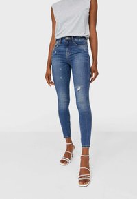 Mujer con jeans ajustados de mezcla azul desgastada con pequeños desgastes y sandalias de tacón alto blancas, combinados con una blusa de tirantes gris claro.