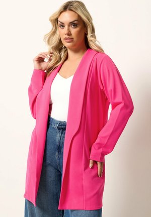 Blond vrouw met een felroze open jas over een wit shirt en blauwe hoge taille jeans, staand tegen een effen achtergrond.