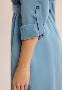 Niebieski strój z podwiniętymi mankietami, z detalem guzika. Gładka tkanina o lekkim połysku, dopasowany fason podkreślający talię.