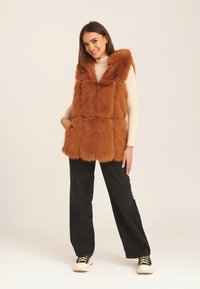 Brown faux fur vest with a hood, featuring a quilted design, front pockets, worn over a beige top and black wide-leg pants.