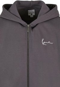 Grå zip-up hettegenser laget av lett stoff, med frontglidelås og hette. Har en liten hvit logo på venstre brystområde. Størrelse L.