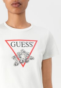 Balta marškinėliai su apvaliu kaklu, su raudonu trikampiu ir "GUESS" logotipu, papuošti gėlių dizainu iš blizgučių ir karoliukų.