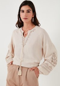 Beige blúz, lekerekített nyakkivágással, hímzett virágmintákkal az ujjakon, és rojt részlettel a nyaknál. Hosszú, laza illeszkedésű ujjak.
