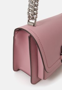 Růžová kožená kabelka se strukturovaným tvarem, zakřiveným klopem, kontrastními tmavými okraji a stříbrným řetízkem na nošení přes rameno. Obsahuje viditelné švy.