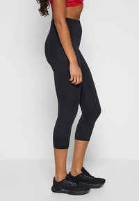 Czarne, wysokie legginsy 3/4 wykonane z rozciągliwego materiału, charakteryzujące się gładkim wykończeniem i dopasowanym krojem, w zestawieniu z czarnymi butami sportowymi.