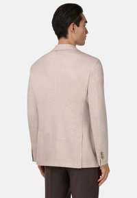 Blazer beige clair avec une texture lisse, design à un bouton, revers en pointe et ornements en bouton sur les manches.