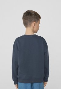 Námořnický modrý svetr s kulatým výstřihem, spadlými rameny a žebrovanými manžetami. Měkká látka s hladkou strukturou. Nosený s modrými džíny.