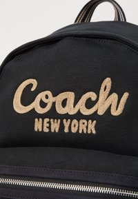Fekete vászon hátizsák hímzett "Coach New York" logóval bézs színben, textúrázott felülettel és fémes cipzár részletekkel.