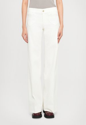Woman wearing high-waist white wide-leg pants with heart-shaped front pockets and dark burgundy platform shoes against a white background.