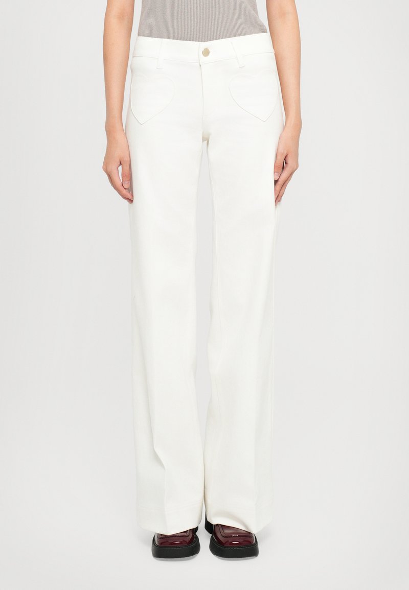 Femme portant un pantalon large taille haute blanc avec des poches frontales en forme de cœur et des chaussures à plateforme bordeaux foncé sur fond blanc.