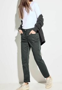 Pantalones de mezcla de algodón verdes con un diseño de pierna recta, bolsillos frontales y cierre de botón, combinados con una blusa blanca de manga larga y zapatillas beige.