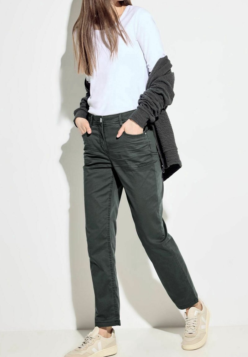 Pantalones de mezcla de algodón verdes con un diseño de pierna recta, bolsillos frontales y cierre de botón, combinados con una blusa blanca de manga larga y zapatillas beige.