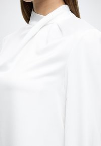 Bluză albă cu guler înalt, textură netedă și drapaj moale. Include pliuri delicate la nivelul gâtului și mâneci lungi.