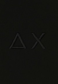 Tessuto nero con simboli ricamati in rilievo neri: un triangolo (Δ) e una X, impostati su una texture a coste verticali.