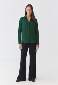 Camicia verde a collare realizzata in tessuto morbido, con bottoni sul davanti, maniche lunghe, abbinata a pantaloni neri a gamba larga e scarpe nere.