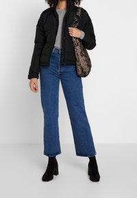 Schwarze Steppjacke mit Reißverschluss, graues Langarmoberteil, blaue weite Jeans und schwarze Wildleder-Stiefeletten mit Absatz. Tasche im Schlangenlederdesign.