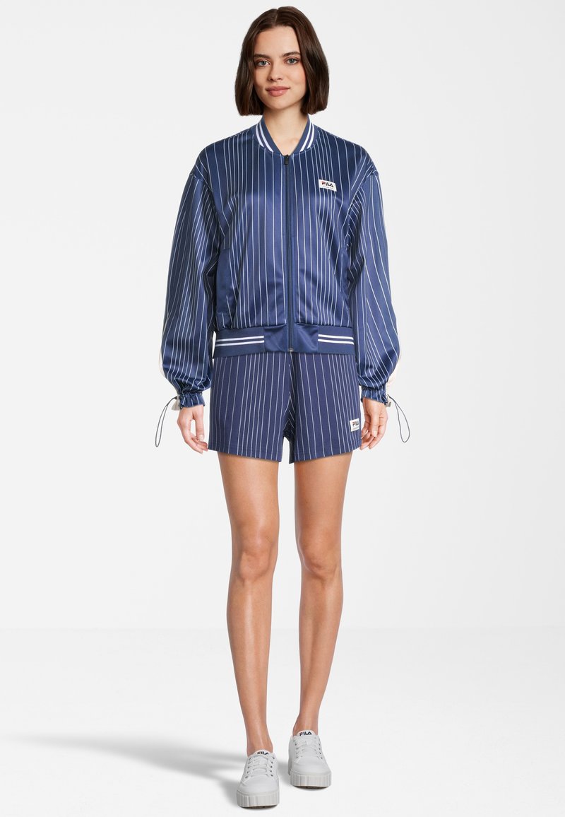 Giacca bomber in poliestere blu navy con strisce verticali bianche, polsini a coste, pantaloni corti abbinati e un piccolo logo applicato, abbinati a sneakers bianche.