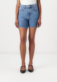 Høyt livd denimshorts i lys blå med klassisk fem-lomme-design og oppbrettede kanter. Kombinert med svarte flate sko.