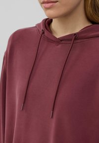Mujer con un hoodie burdeos con cordones, mostrando el área del cuello y los hombros, con tono de piel natural y labios cerrados.