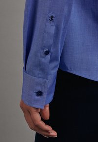 Blue striped shirt cuff with two dark buttons and an embroidered rose detail. Made of a smooth fabric, showcasing a tailored design.