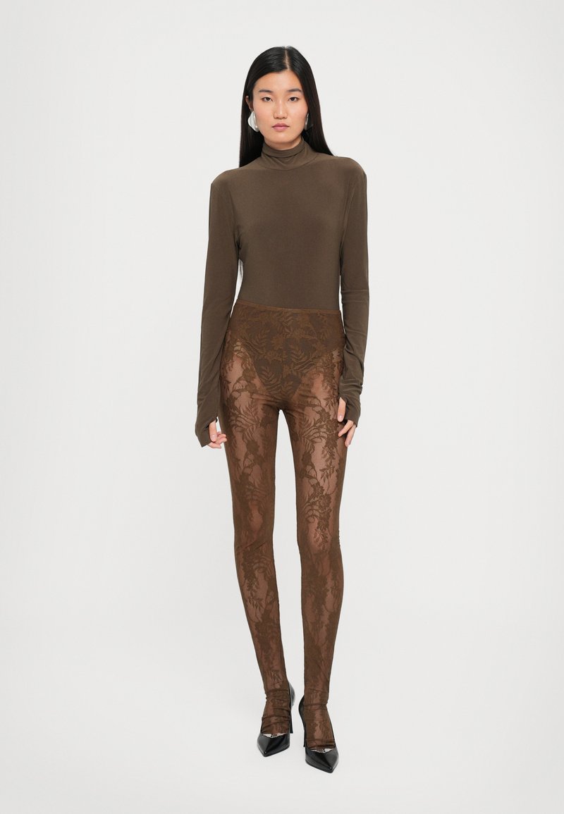 Braunes Langarmoberteil kombiniert mit halbdurchsichtigen braunen Blumen-Lace-Leggings. Enganliegendes Design mit aufwändigem Blattmuster und konturierender Passform. Schwarze Absatzschuhe.