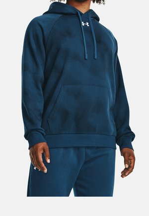 Personne portant un sweat à capuche bleu foncé texturé avec une poche à l'avant et un pantalon bleu assorti, debout avec les bras détendus le long du corps.