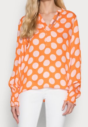 Kvinde iført en langærmet orange bluse med store lyserøde polkaprikker og hvide bukser, vist fra nakke til hofter.