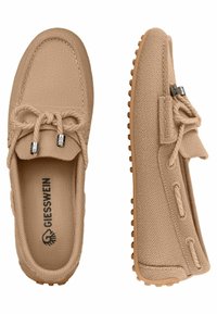 Beige Strick-Slipper mit strukturiertem Obermaterial, flexibler Gummisohle, dekorativen Schnürsenkeln und Logo auf der Innensohle. Die Seitenansicht zeigt das weiche Material.