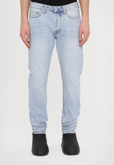 Világoskék farmernadrág, egyenes szabással, öt zsebbel és diszkrét fakulással. Fekete slip-on cipővel párosítva.