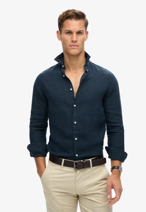 Navyblauwe linnen overhemd met een hoge kraag, voorzien van een knoopsluiting aan de voorkant en opgerolde manchetten. G gedragen met beige broeken en een zwarte riem.