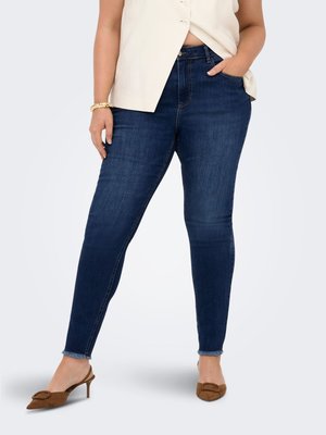 Kropsformet person iført mørkeblå skinny jeans med frynsete kanter, beige ærmeløs top, guldbøjle og brune spidse kitten heels.
