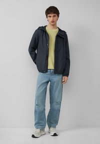 Donker marineblauwe lichtgewicht jas met capuchon, lichtgele geribde trui, versleten lichtblauwe spijkerbroeken en witte sneakers. Casual pasvorm overal.