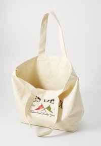 Cotton tote bag in cream with black text and colored flag motifs; wide opening, two long handles, and a soft, textured finish.